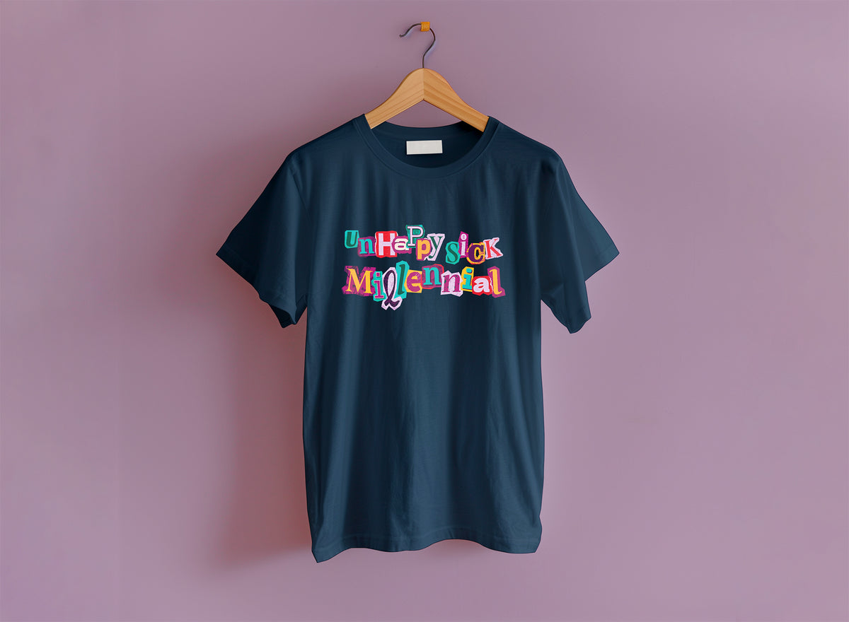 A navy Comfort Colors short-sleeve t-shirt hanging on a wooden hanger against a solid lavender wall. The shirt features a colorful graphic text print on the chest reading 'unhappy sick millennial' in a multicolored, layered font.